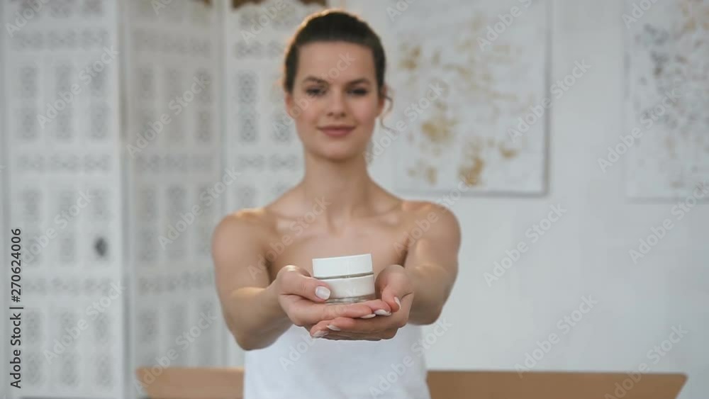 Handsome cute woman stretching out jar with cream to the camera on the bathroom backgound. Handsome cute woman stretching out jar with cream to the camera on the bathroom backgound.
