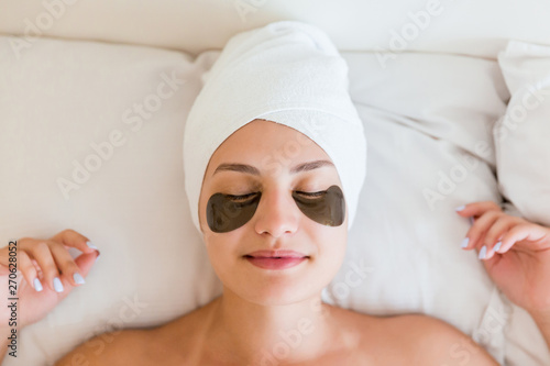 Beautiful young woman with under eye patches in bathrobe lying in bed. Happy girl taking care of herself. Beauty skincare and wellness morning concept