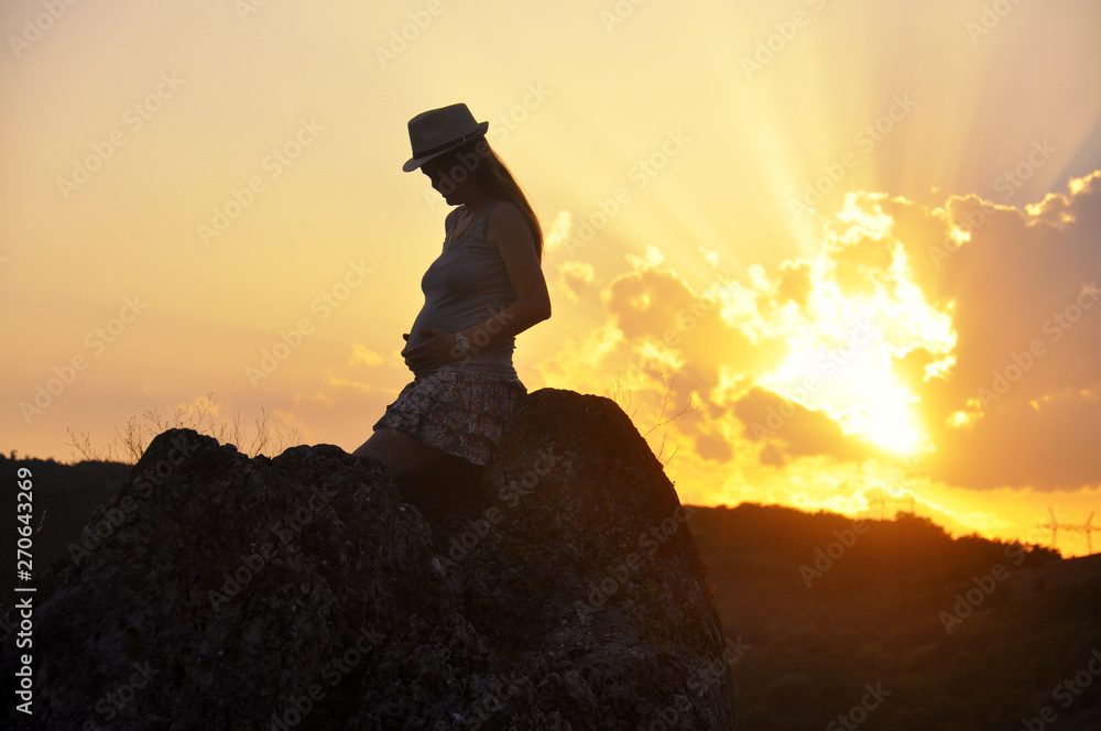 Pregnant woman with hat look her belly and enjoy in sunset on mountain. Pregnant woman on top of mountain at sunset