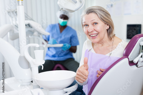 Happy cute mature woman sitting in dental chair after teeth cure giving thumb up © JackF