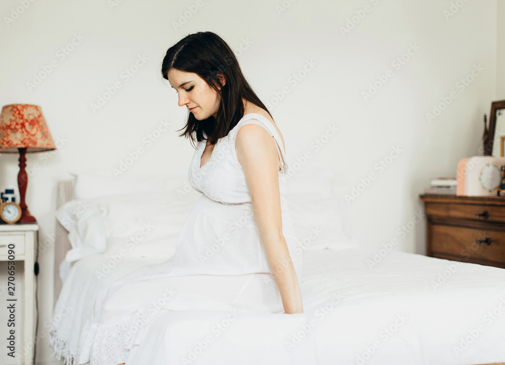 Thoughtful expectant woman sits on bed in bright bedroom Thoughtful expectant woman sits on bed in bright bedroom