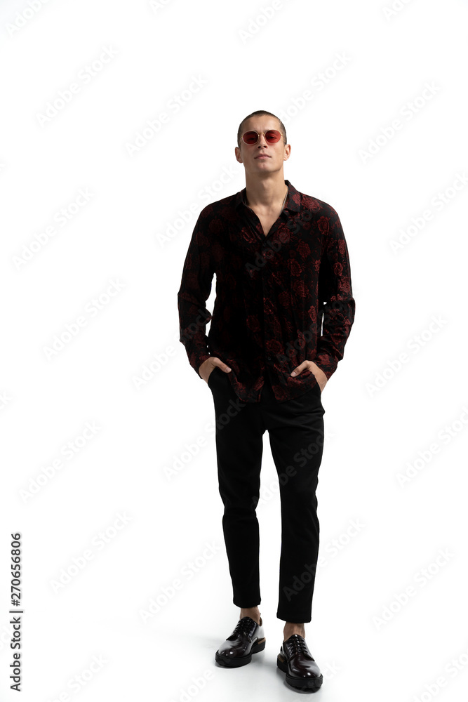 Fototapeta premium Full length portrait of amazing attractive man in stylish trendy clothes, wearing red sunglasses, holding hands in pockets, looking at camera.