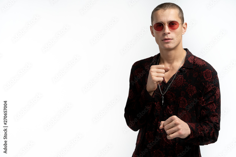 Attractive young man in trendy stylish black shirt with rose print in red sunglasses keeps fists in front if him, looking at the camera, isolated over white background Attractive young man in trendy stylish black shirt with rose print in red sunglasses keeps fists in front if him, looking at the camera, isolated over white background