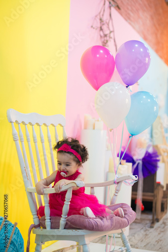 Happy smiling sweet baby girl sitting on armchair with birthday balloons.