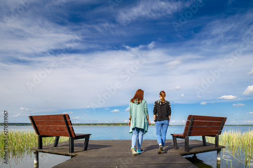 Two young girls girlfriends walk in the fresh air on the pier of the reservoir in beautiful weather on a Sunny spring day. People in solitude with nature. Walking in the fresh air as way of lifestyle. © ruslan_khismatov