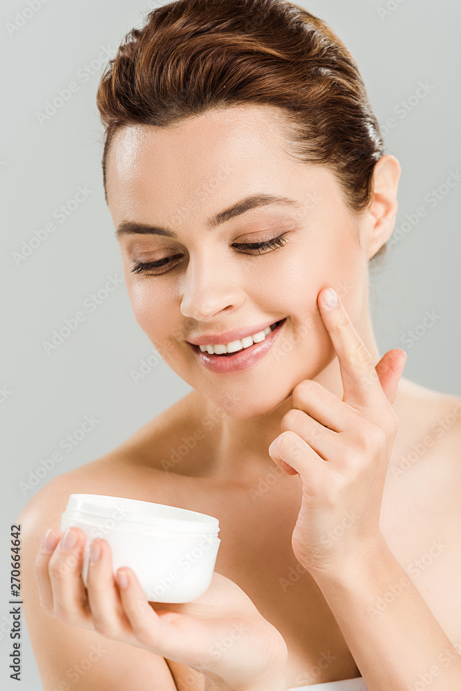 happy woman looking at container while applying face cream isolated on grey happy woman looking at container while applying face cream isolated on grey