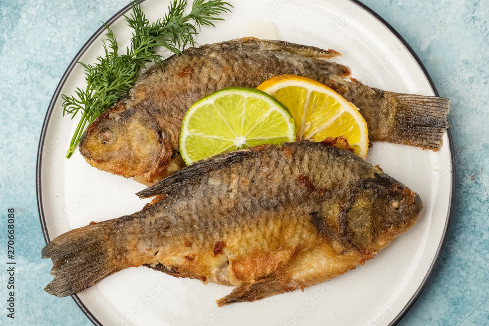 Fried fish Crucian on a plate with lemon and lime slices, top view. Ready meal. Blue background Fried fish Crucian on a plate with lemon and lime slices, top view. Ready meal. Blue background