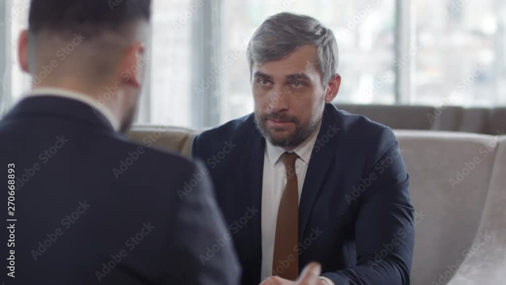 Rear view of middle-aged gray-headed businessman sitting at table, listening carefully to his business partner sitting in front of him while reading documents lying on table Rear view of middle-aged gray-headed businessman sitting at table, listening carefully to his business partner sitting in front of him while reading documents lying on table