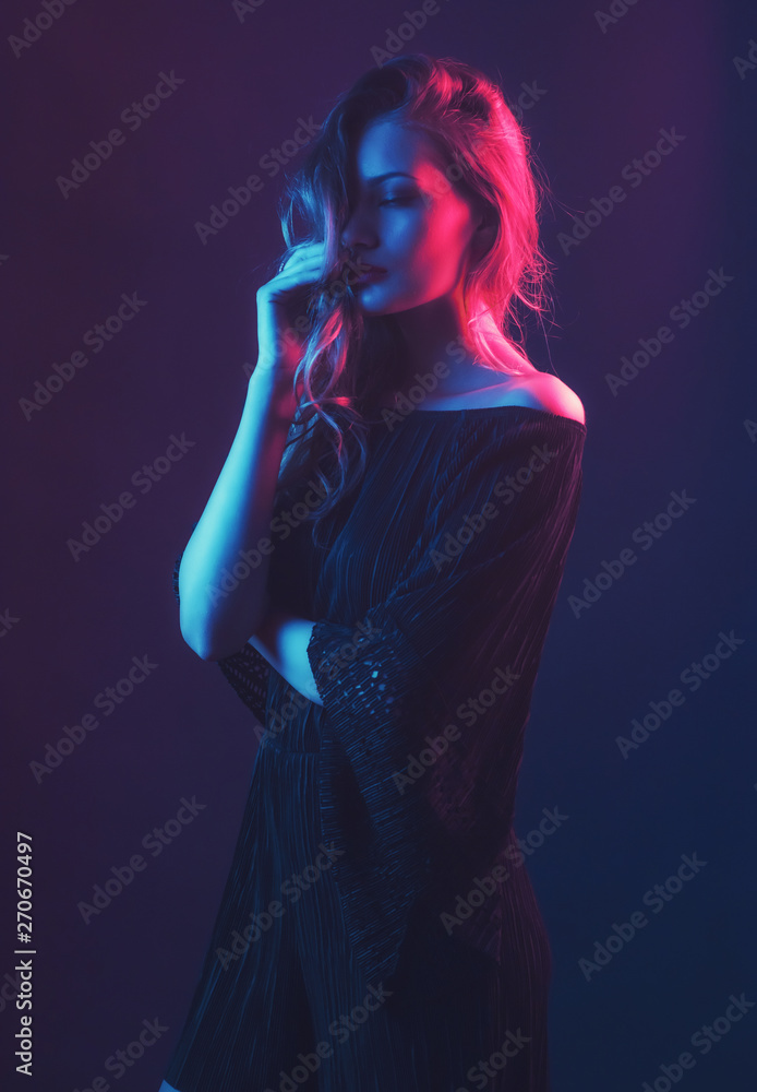 Beautiful ,young blonde with bright scarlet lips and expressive eyes in a black jumpsuit in neon light blue and red lamps.