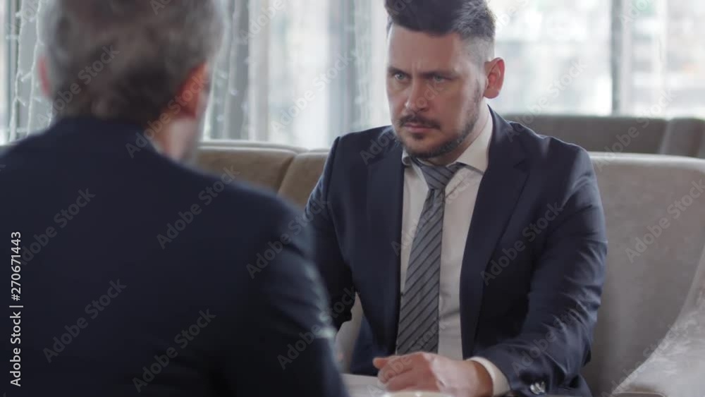 Back view of black-haired office worker in suit sitting on sofa at café table, listening to his colleague sitting in front of him while using his smartphone Back view of black-haired office worker in suit sitting on sofa at café table, listening to his colleague sitting in front of him while using his smartphone