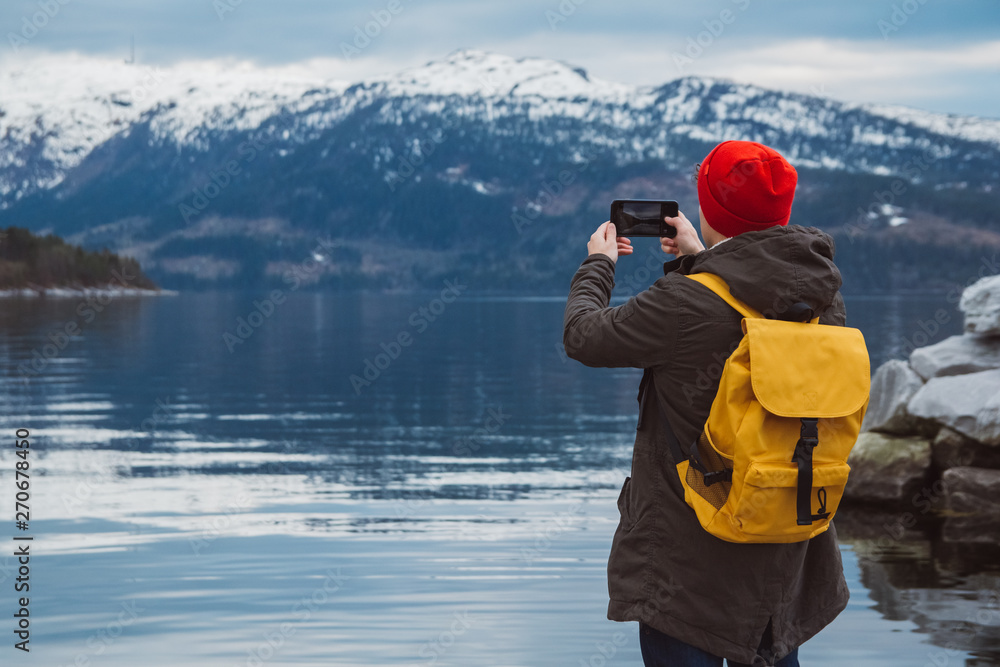 Traveler man taking photo with smartphone on mountains. Tourist in yellow backpack standing on a background of a mountain and a lake. Traveler walks, takes photo in mountains. View from back.