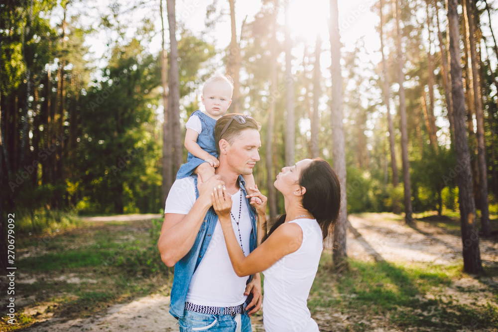Fototapeta premium Family mom, dad and daughter sits at the daddy on the shoulders, and parents kiss on the nature in the forest in the summer at sunset