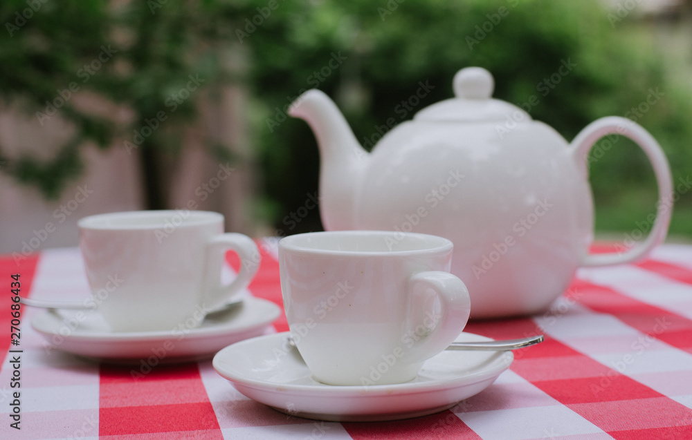 White tea service for two. Tea pair with a teapot and a milk jug on a bright multi-colored checkered tablecloth White tea service for two. Tea pair with a teapot and a milk jug on a bright multi-colored checkered tablecloth