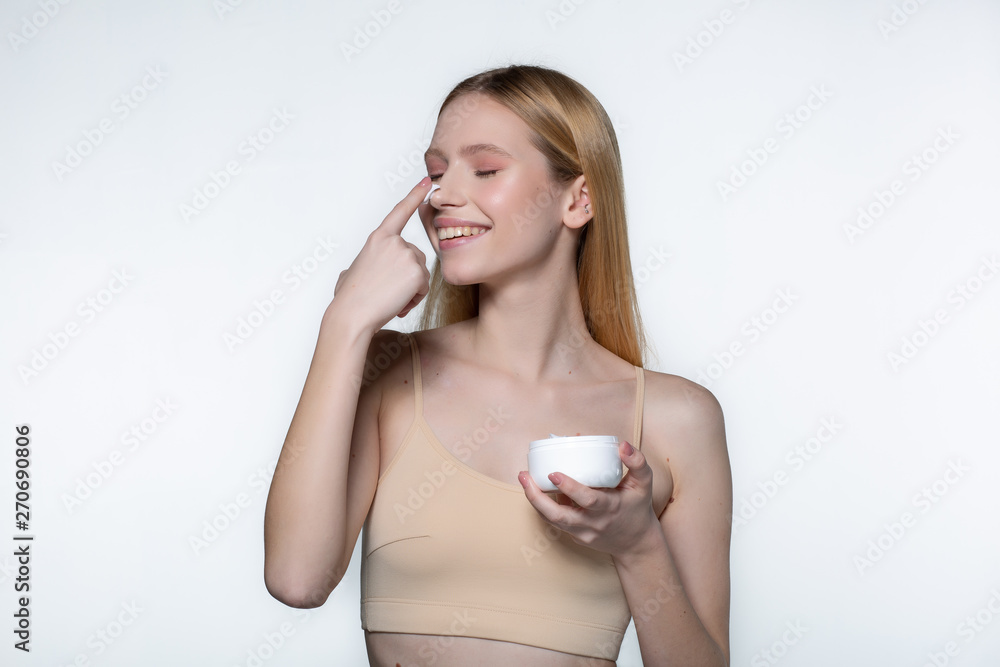 Close up of positive young woman with blonde hair standing with a jar of cream in her hand and smiling while putting small amount of cream on her nose. Close up of positive young woman with blonde hair standing with a jar of cream in her hand and smiling while putting small amount of cream on her nose.