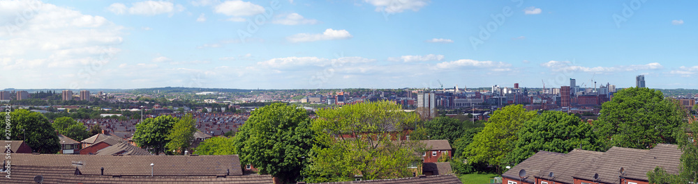 a wide panoramic view showing the whole of leeds city center with towers apartments roads and commercial buildings surrounded by trees and roofs against a blue sky