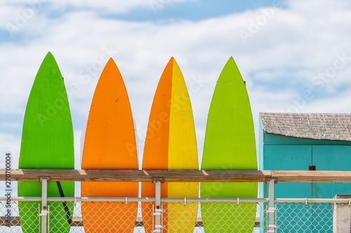 Stack row of multicolored colorful stand up surfing boards on railing fence with green orange and yellow color against blue sky lifeguard tower or rental shop photo