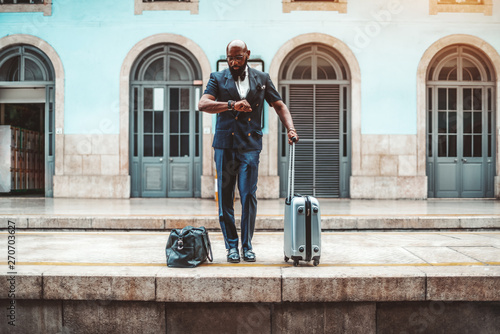 A handsome bald bearded African male entrepreneur in a fashion vest and glasses is waiting for his train and looking at his watch on the railway platform to go on a business trip with two travel bags