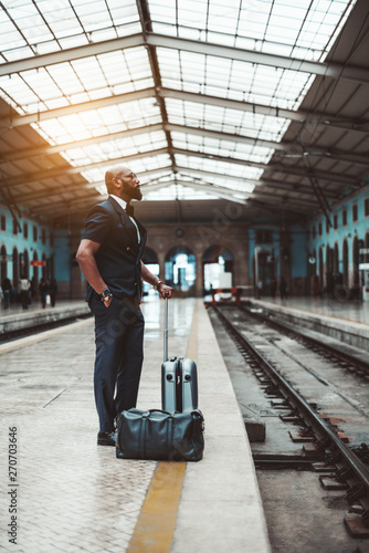 A fancy black guy with travel bags is waiting for his train on the platform of a railway station depot; vertical shot of an African businessman waiting for the train for his business trip indoors
