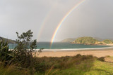 The end of a double rainbow over the deserted, sandy beach at Matai Bay with a small, silhouetted bush and flax in the foreground, Karikari Peninsula, Northland, New Zealand.