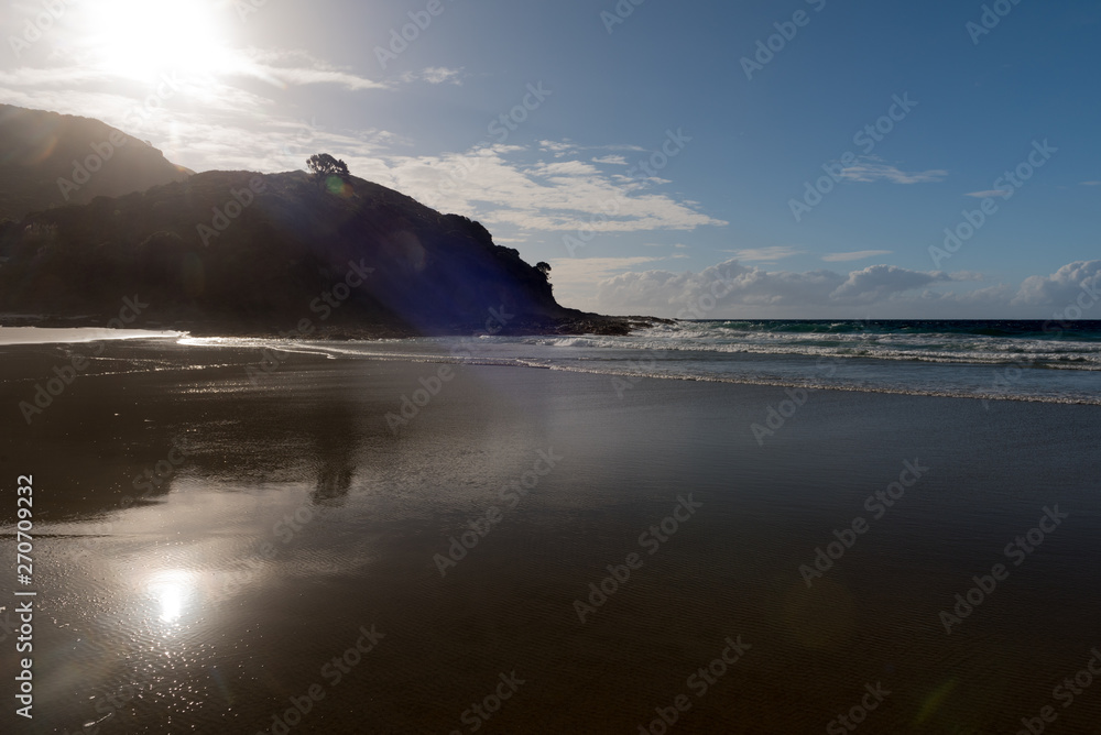 The backlit, silhouetted cliffs and headland at the end of Matai Bay reflected in the wide, sandy beach. Karikari Peninsula, Northland, New Zealand. The backlit, silhouetted cliffs and headland at the end of Matai Bay reflected in the wide, sandy beach. Karikari Peninsula, Northland, New Zealand.