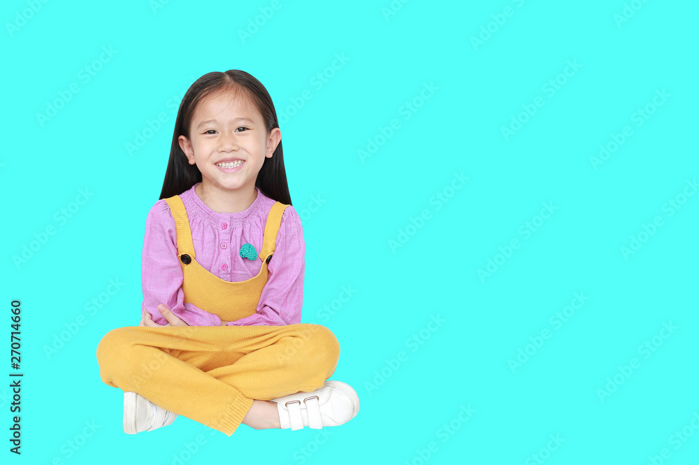 Portrait of happy little Asian child girl in pink-yellow pink-yellow dungarees sitting isolated over cyan background with copy space. Portrait of happy little Asian child girl in pink-yellow pink-yellow dungarees sitting isolated over cyan background with copy space.