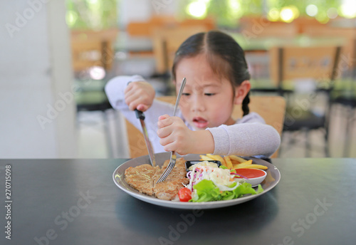 Asian child girl eating Pork steak and vegetable salad on the table with holding knife and fork. Children having breakfast.