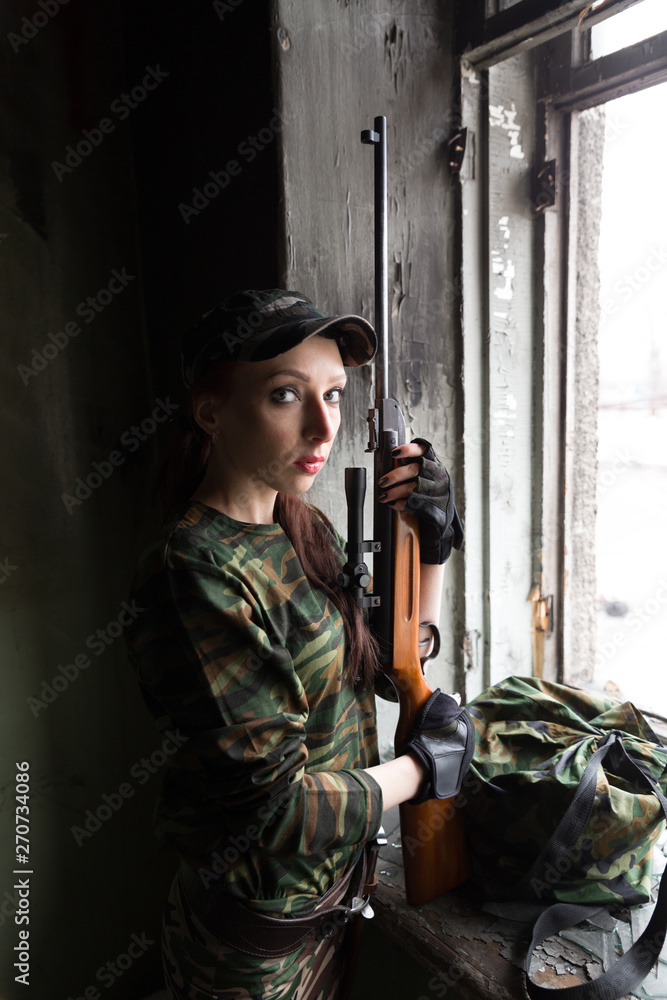 A thin red-haired girl stands at the broken window with a rifle. The girl in green camouflage with a gun. Military service for girls. The girl is in the army. A thin red-haired girl stands at the broken window with a rifle. The girl in green camouflage with a gun. Military service for girls. The girl is in the army.