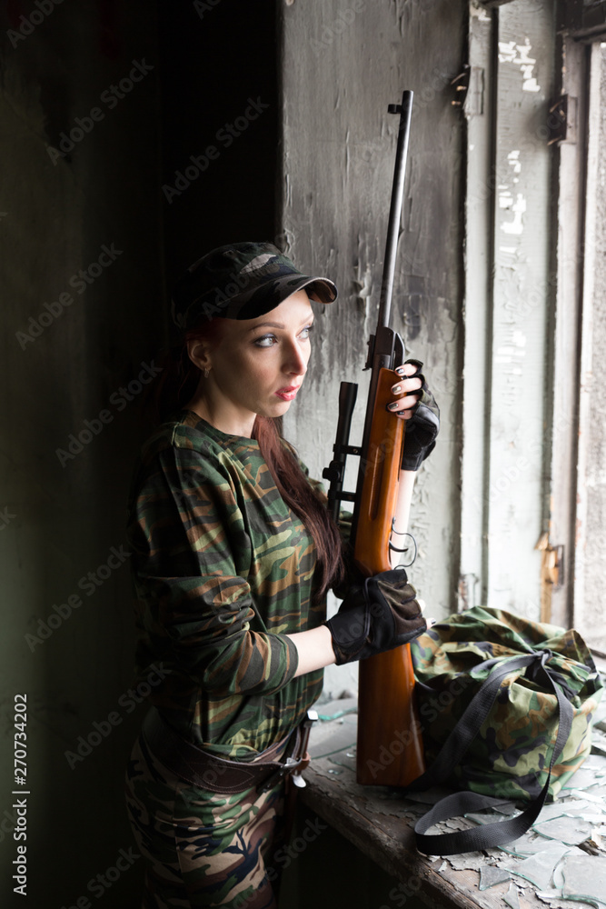 A thin red-haired girl stands at the broken window with a rifle. The girl in green camouflage with a gun. Military service for girls. The girl is in the army.