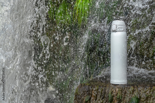 White thermos of tea or coffee on stone near waterfall with splashes of water drops. Adventure hiking tourism concept.