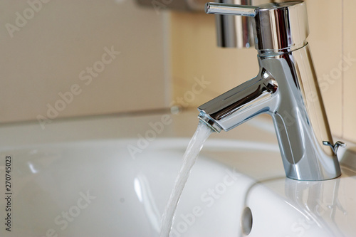 Water from the tap. A stream of clean water flows into the sink. Open chrome faucet washbasin. Warm tones.