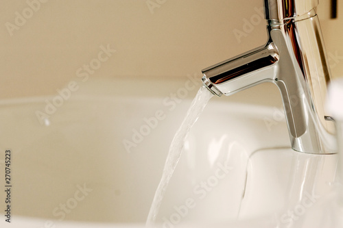 Water from the tap. A stream of clean water flows into the sink. Open chrome faucet washbasin. Warm tones.