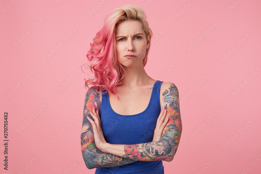 Young sad frowning beauty woman with pink hair, stands with crossed arms over pink background, looks displeased, wears a blue shirt. People and emotion concept. Young sad frowning beauty woman with pink hair, stands with crossed arms over pink background, looks displeased, wears a blue shirt. People and emotion concept.