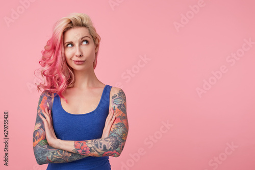 Portrait of young thinking cute lady with pink hair, wears blue shirt, stands over pink background copy space with crossed arms, looks up thoughtfully. People and emoyion concept. photo