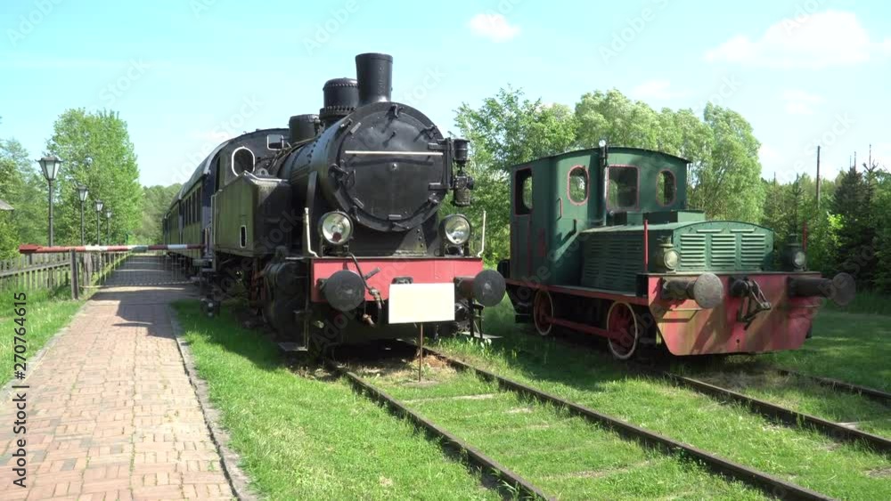 Two old trains are standing at the station. One black, red locomotive and green, red. Retro steam train on railway station in summer. Two old trains are standing at the station. One black, red locomotive and green, red. Retro steam train on railway station in summer.