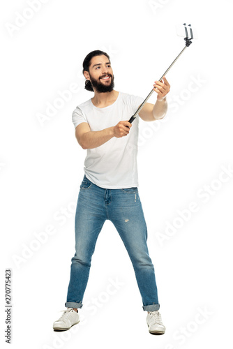 happy latin man holding selfie stick and taking selfie while standing isolated on white