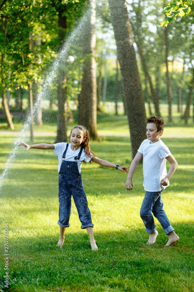 Cheerful little girl and boy playing near watering on green grass and smiling. Brother and sister in wet clothes in the summer park. Best friends. Childhood concept.