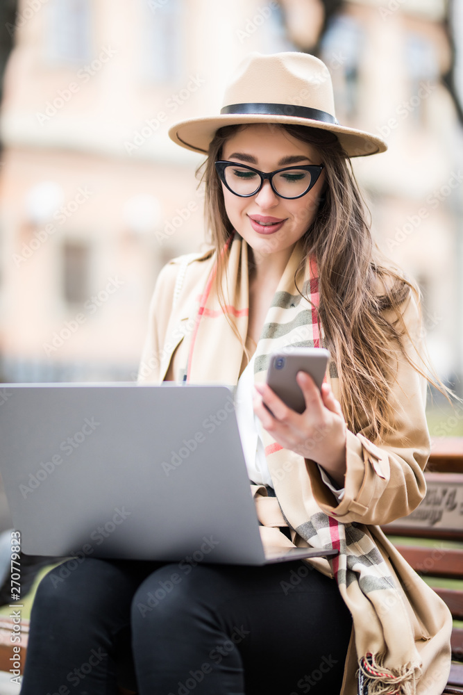 Smiling young woman sitting on bench outdoors at the street, using laptop computer, use mobile phone Smiling young woman sitting on bench outdoors at the street, using laptop computer, use mobile phone