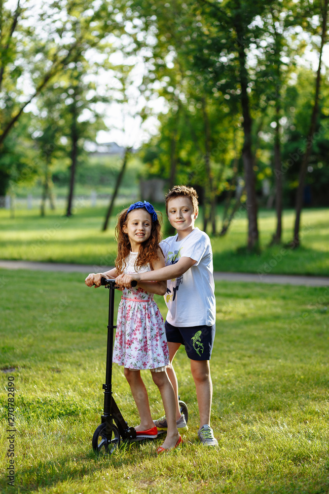 Fototapeta premium Cheerful little girl and boy drive the scooter on green grass. Brother and sister playing in the summer park. Best friends. Childhood concept.