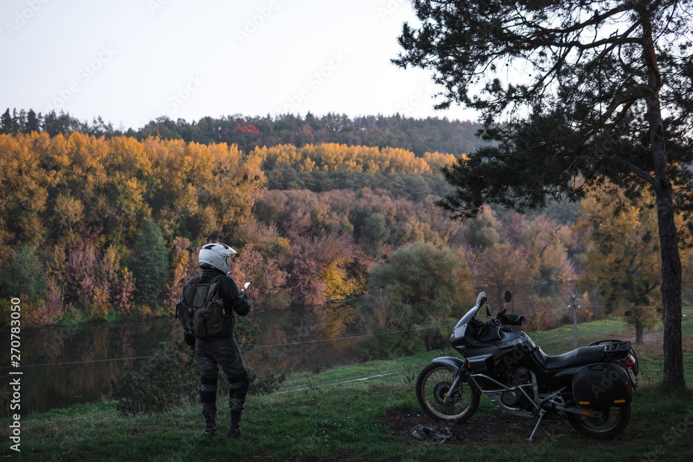 Obraz premium Rider standing with adventure motorcycle, Motorcyclist gear, A motorbike driver looks, active lifestyle, enduro travel road trip. Evening, autumn. Tourist traveler. copy space, use gadget, enternet