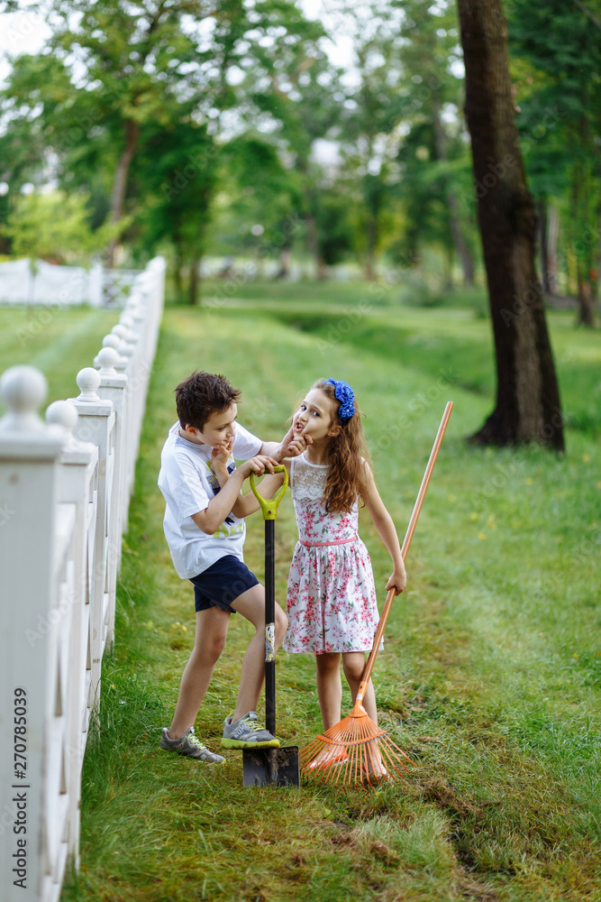 Fototapeta premium Cheerful little girl with rake and boy digging with shovel near the wooden fence in the park. Brother and sister works in garden. Childhood concept.
