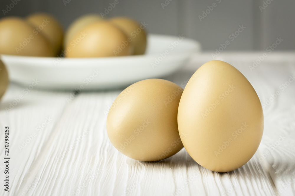 Two pheasant eggs in the foreground and a plate with eggs in the background on a wooden white table Two pheasant eggs in the foreground and a plate with eggs in the background on a wooden white table