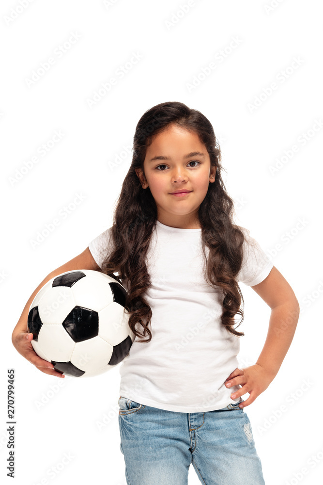 cheerful latin kid standing with hand in hip and holding soccer ball isolated on white