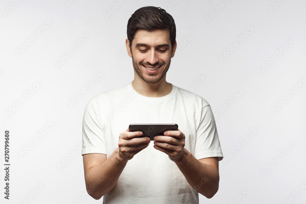 Handsome young man in white t-shirt, holding smartphone, playing video game app, isolated on gray background Handsome young man in white t-shirt, holding smartphone, playing video game app, isolated on gray background