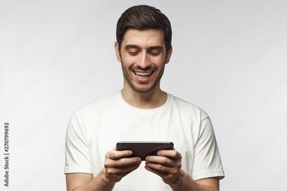 Handsome laughing young man in white t-shirt, holding smartphone, playing video game, isolated on gray background Handsome laughing young man in white t-shirt, holding smartphone, playing video game, isolated on gray background
