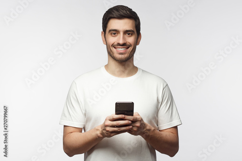 Handsome smiling man looking at camera while holding his smart phone, isolated on gray background © Damir Khabirov