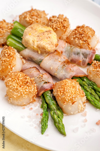 Fried seared scallops breaded with sesame. Garnished with fried green asparagus and bacon. Large portion on a white plate. Close up and vertical view. .