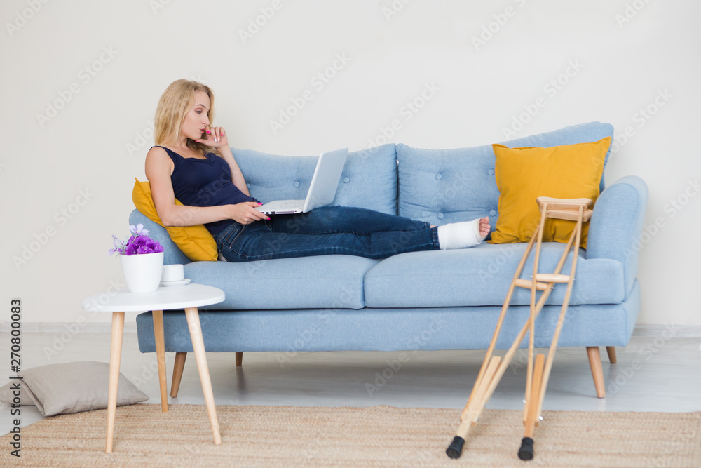 Young woman with broken leg in cast sitting on sofa at home working on the laptop Young woman with broken leg in cast sitting on sofa at home working on the laptop