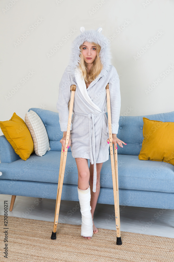 Φωτογραφία Young woman in a dressing gown with crutch and broken leg in cast in the room