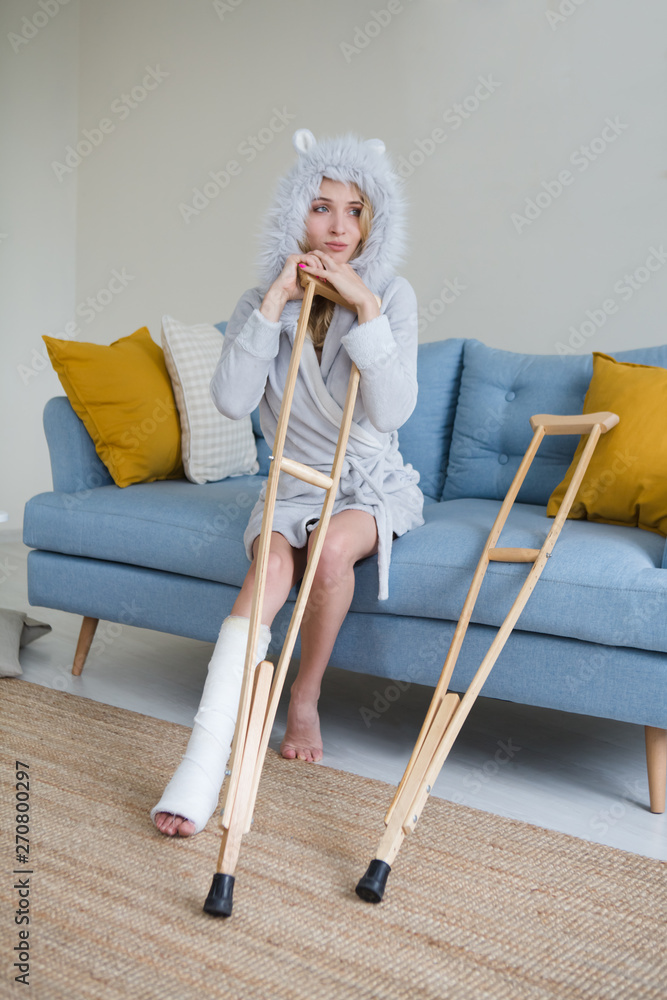 Φωτογραφία Young woman in a dressing gown with crutch and broken leg in cast in the room