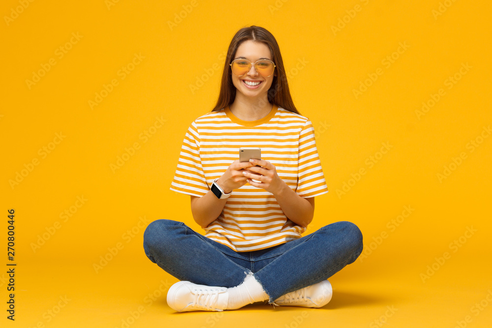 Happy young woman sitting on the floor, holding smartphone in hands and looking at camera, isolated on yellow background Happy young woman sitting on the floor, holding smartphone in hands and looking at camera, isolated on yellow background